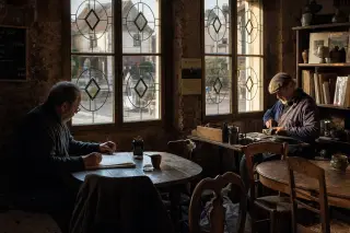Two local artisans quietly working inside an old village café, golden afternoon light streaming through leaded windows onto rustic wooden tables and shelves, with a warm, contemplative atmosphere.