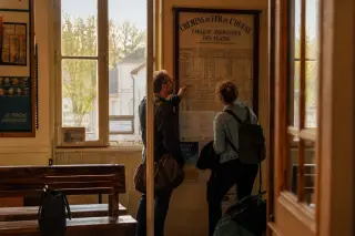 Two people quietly checking a train timetable inside a sunlit rural station, warm afternoon light and worn benches creating a calm, authentic ambiance.