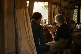 Two people share a quiet, focused conversation in a warmly lit village workshop, surrounded by wood and tools, as golden sunset light filters through a window.