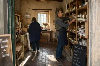 shopkeeper arranging shelves in a village grocery, customer browsing, warm afternoon light through window, rustic Perche interior
