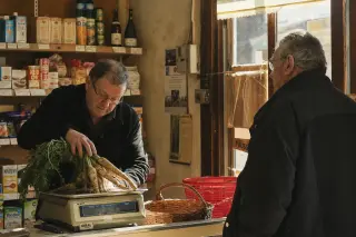 Two people chatting by the counter of a modest rural grocery, afternoon sunlight streaming in, shelves lined with everyday products, in a warm, lived-in ambiance.